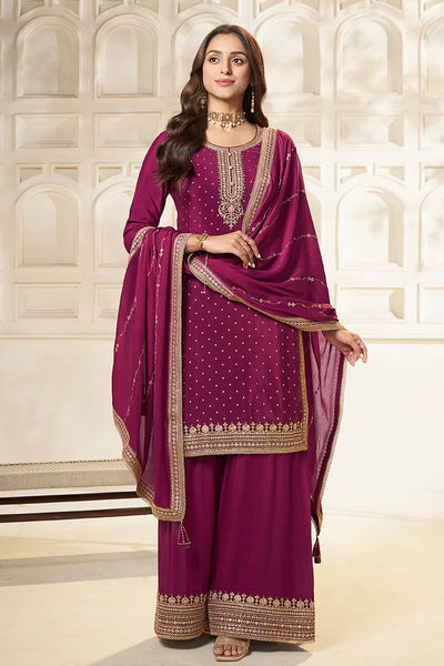 Woman wearing a traditional plum sharara suit outfit with gold embroidery against a white architectural backgroundWoman wearing a traditional plum sharara suit outfit with gold embroidery against a white architectural background