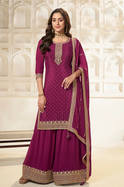 Woman wearing a purple plum sharara suit traditional outfit with gold embroidery against a white background