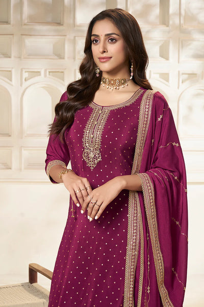 Woman wearing a purple plum sharara suit traditional outfit with gold embroidery against a white wall.Woman wearing a purple plum sharara suit traditional outfit with gold embroidery against a white wall.