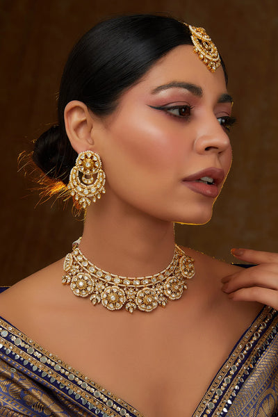 Woman wearing gold kundan jewelry including earrings, necklace, and tikka headpiece against a brown background