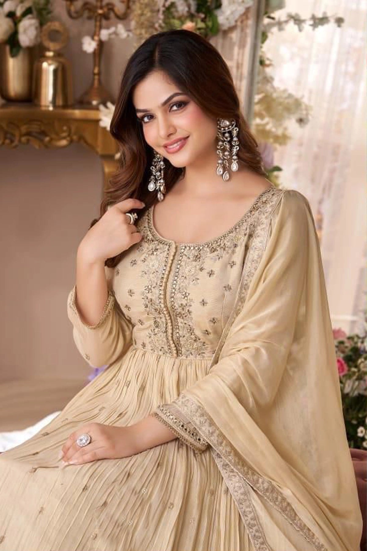Woman wearing a beige embroidered traditional anarkali outfit with jewelry in an elegant indoor setting.