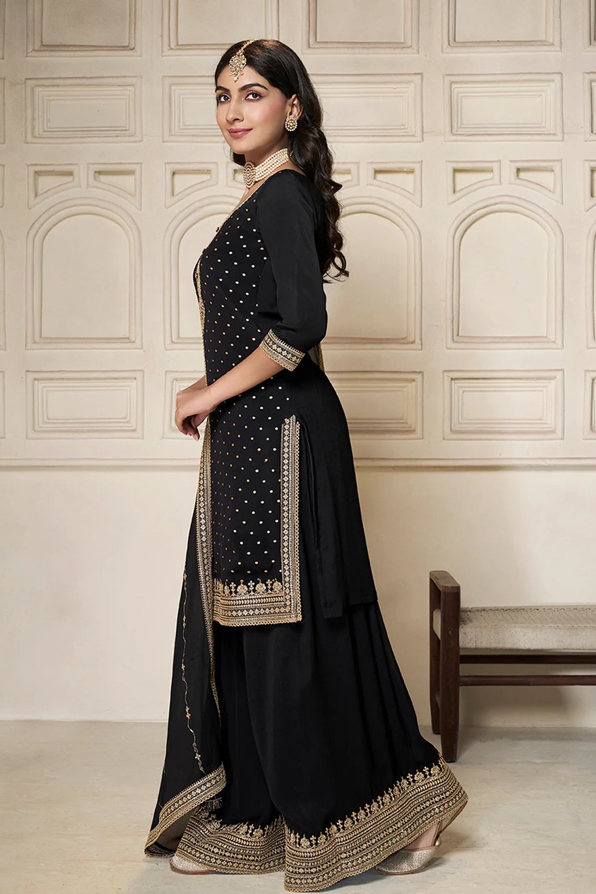 Woman wearing a black sharara suit traditional outfit with gold embroidery against a beige wall.