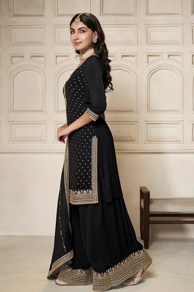 Woman wearing a black sharara suit traditional outfit with gold embroidery against a beige wall.Woman wearing a black sharara suit traditional outfit with gold embroidery against a beige wall.