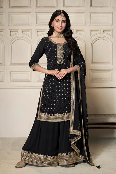Woman wearing a black sharara suit traditional outfit with gold embroidery against a beige wall.