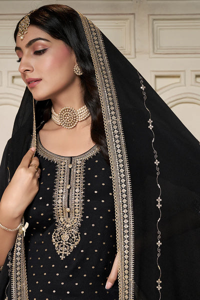 Woman wearing a black sharara suit traditional outfit with gold embroidery and jewelry against a beige wall.Woman wearing a black sharara suit traditional outfit with gold embroidery and jewelry against a beige wall.
