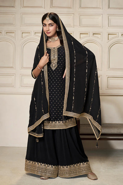 Woman wearing a black sharara suit traditional outfit with gold embroidery against a beige wall.Woman wearing a black sharara suit traditional outfit with gold embroidery against a beige wall.