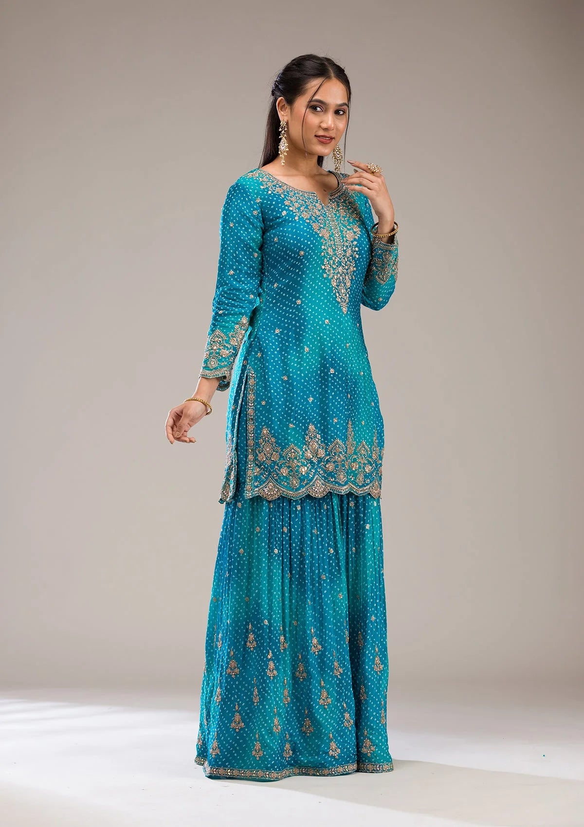 Woman wearing a turquoise embroidered traditional sharara outfit on a plain background