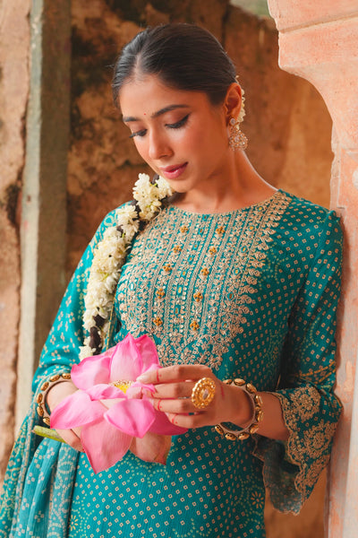 Woman in traditional teal blue attire holding a pink flower, standing against an architectural background.Woman in traditional teal blue attire holding a pink flower, standing against an architectural background.