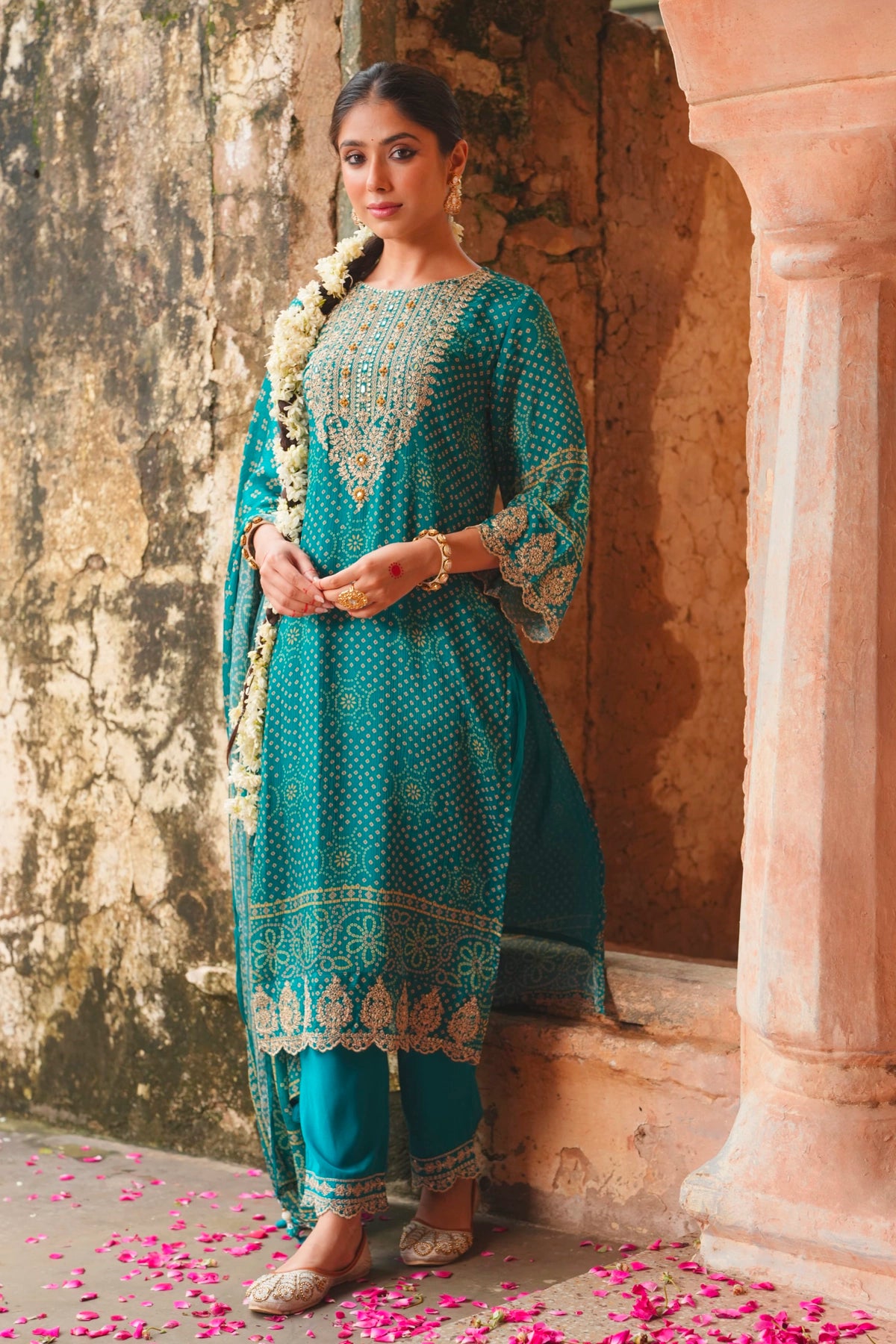 Woman in a teal blue traditional outfit standing in an architectural setting with pink petals on the ground.