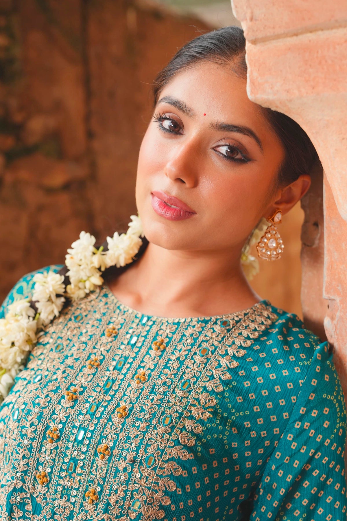 Woman in traditional teal blue attire with floral decorations, standing against a stone wall.