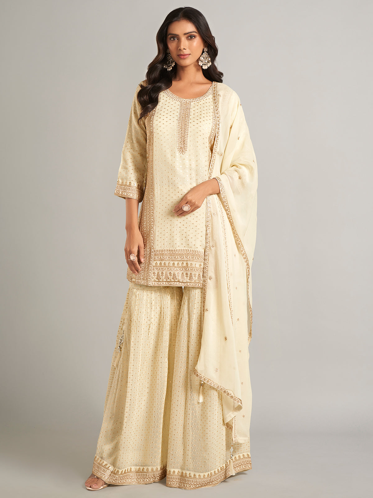 Woman wearing a cream traditional outfit with intricate patterns on a gray background