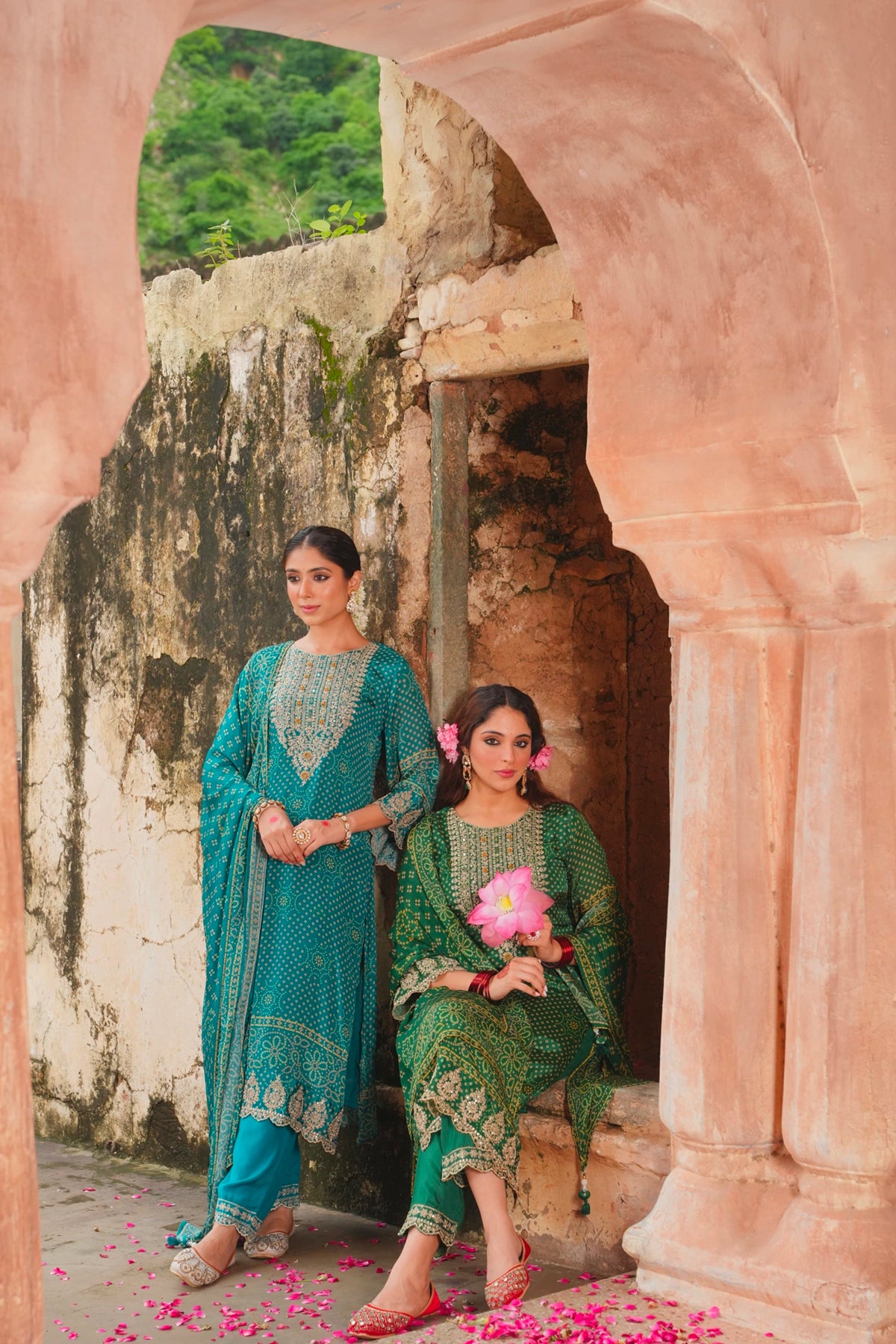 Two women in traditional outfits standing in front of a stone archway with greenery in the background.