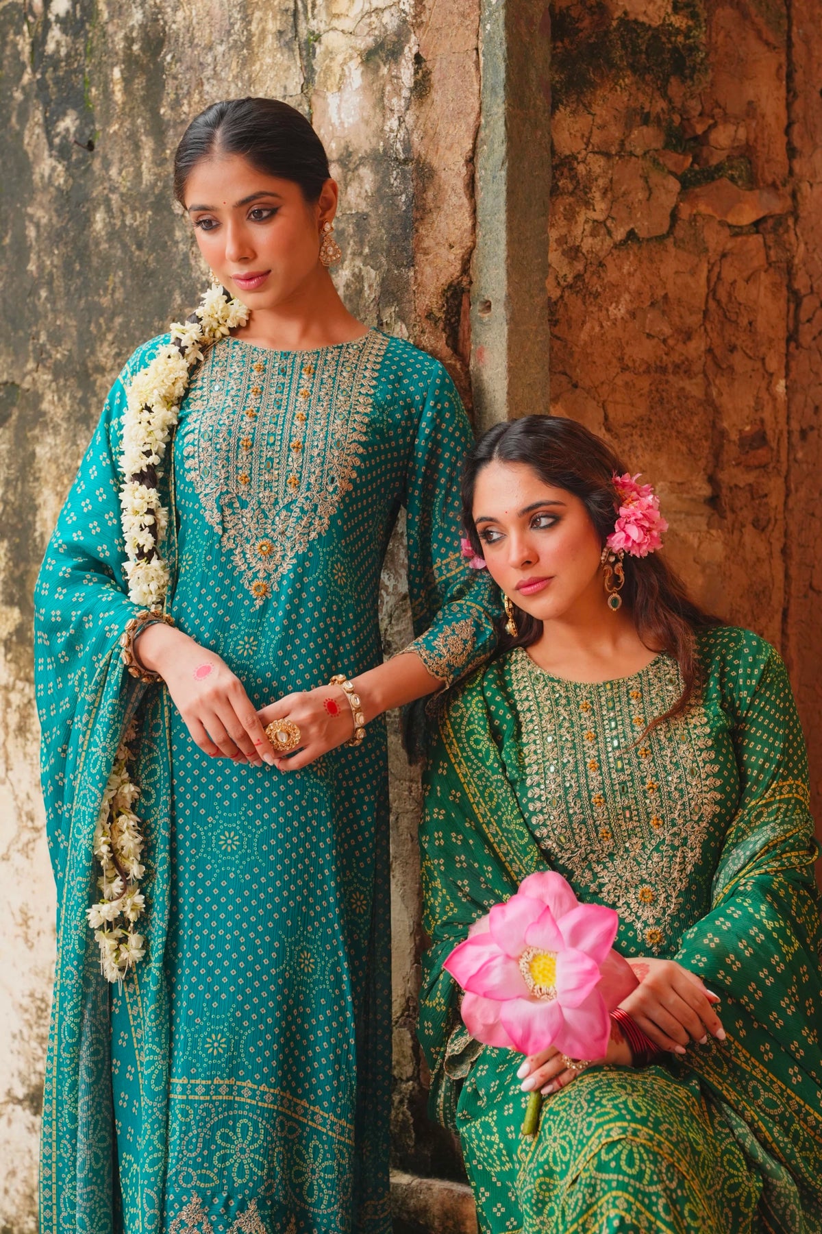 Two women in traditional green and teal outfits with floral accessories against a textured wall.