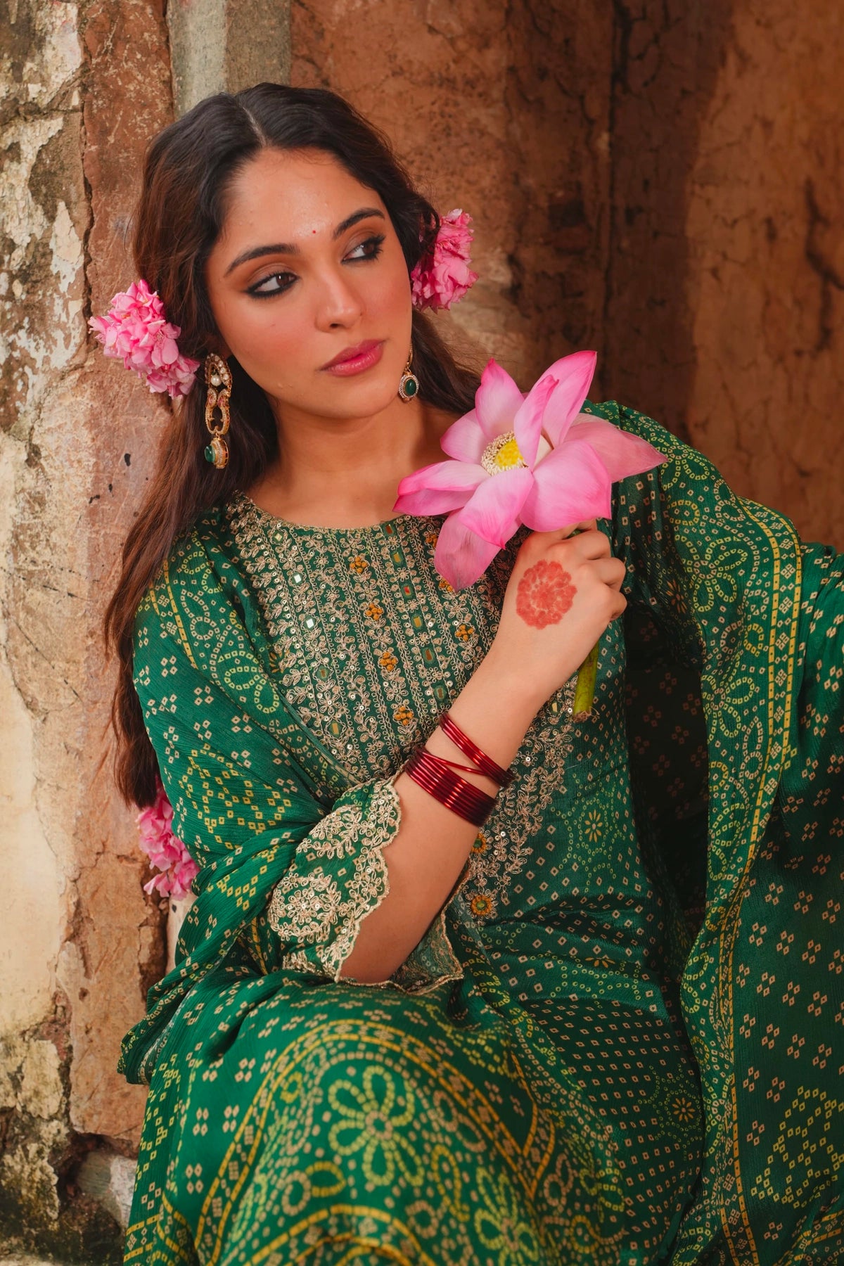 Woman in a green traditional outfit holding pink flowers against a stone wall.