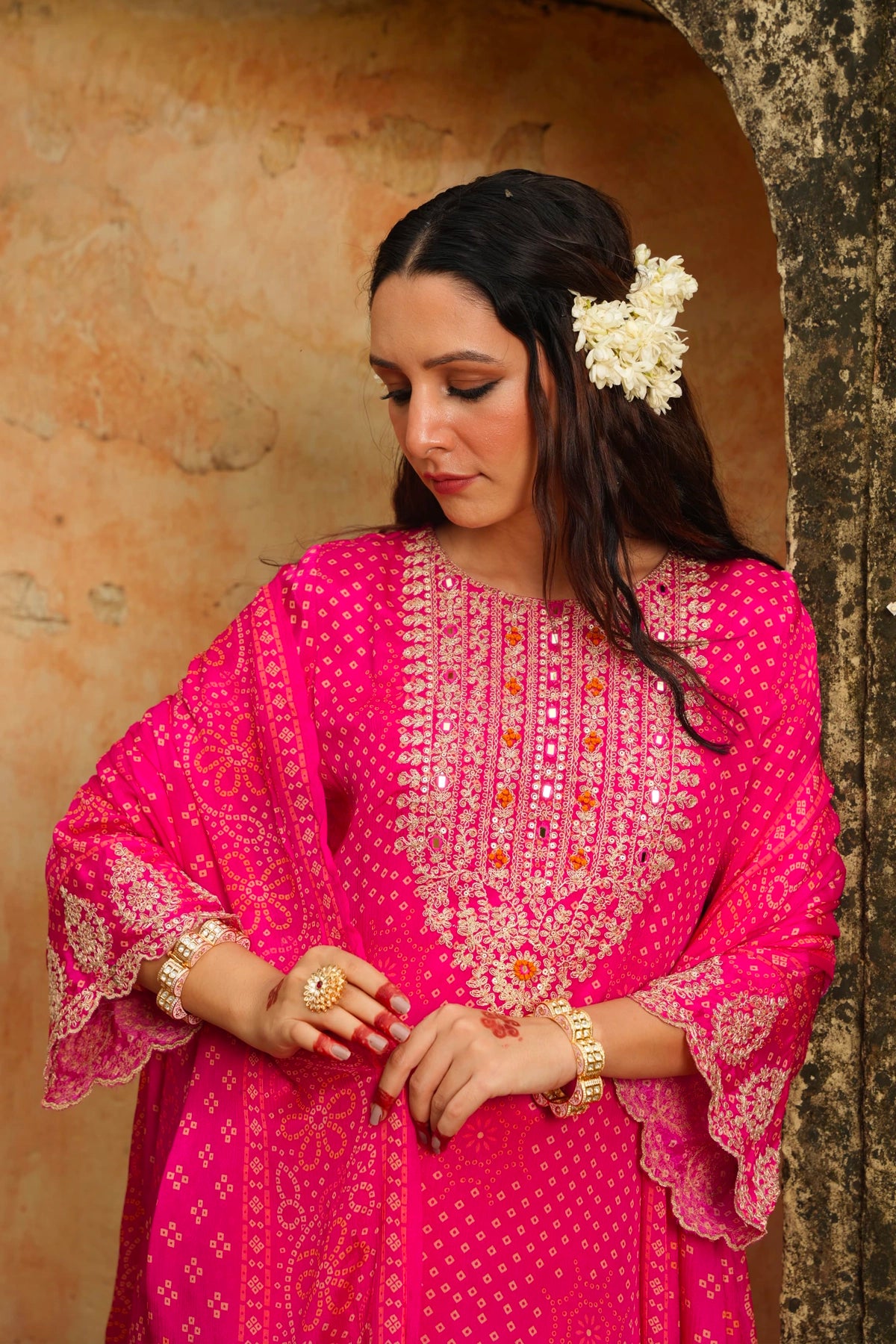 Woman wearing a pink traditional outfit with intricate designs, standing against a textured wall.
