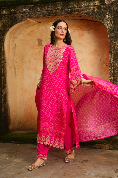 Woman in a bright pink traditional kurta set outfit with gold embroidery standing in front of an archway.