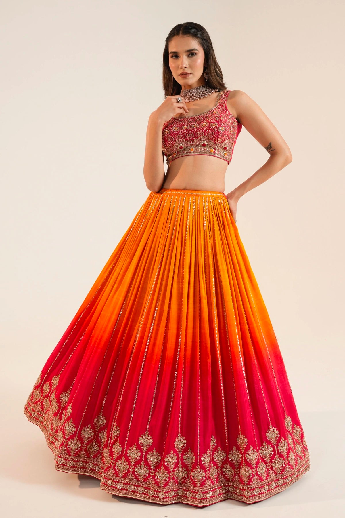 Woman wearing a traditional lehenga choli outfit with a gradient skirt from orange to red on a plain background