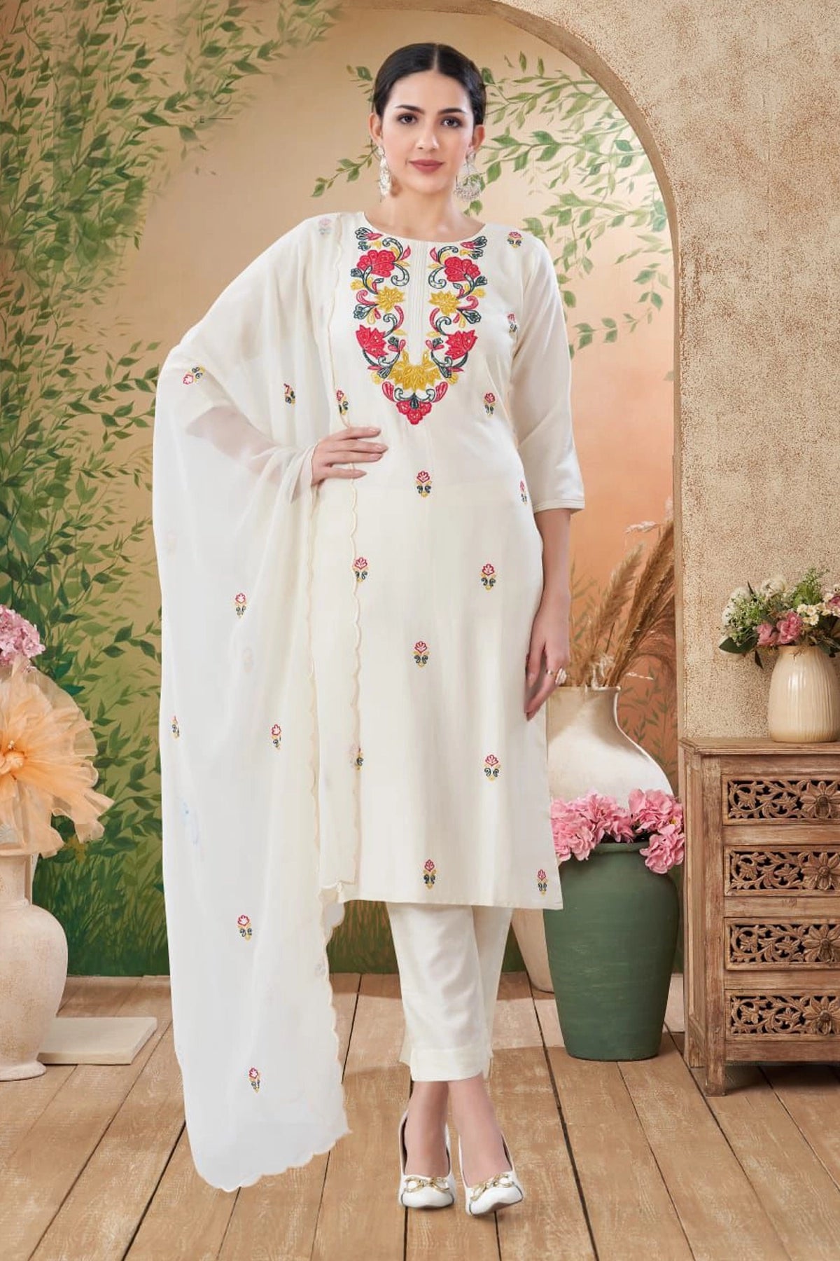 Woman in a white embroidered outfit standing in a decorative indoor setting with plants and flowers.