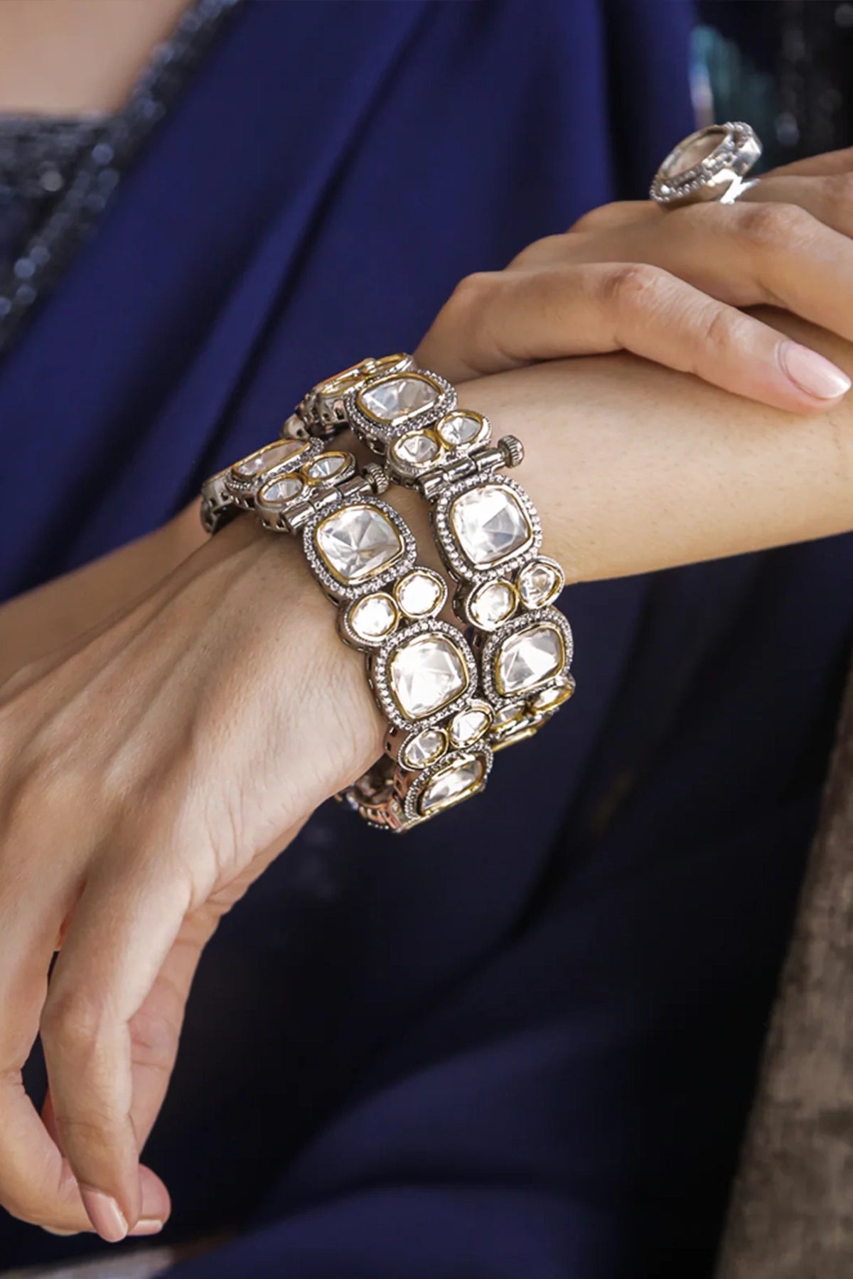 Close-up of a hand wearing a decorative polki kada bracelet with a blue fabric background