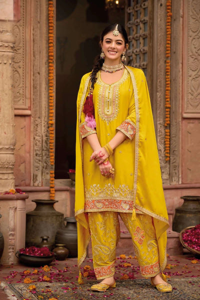 Woman in a yellow traditional punjabi outfit with intricate patterns standing in front of decorative elements.