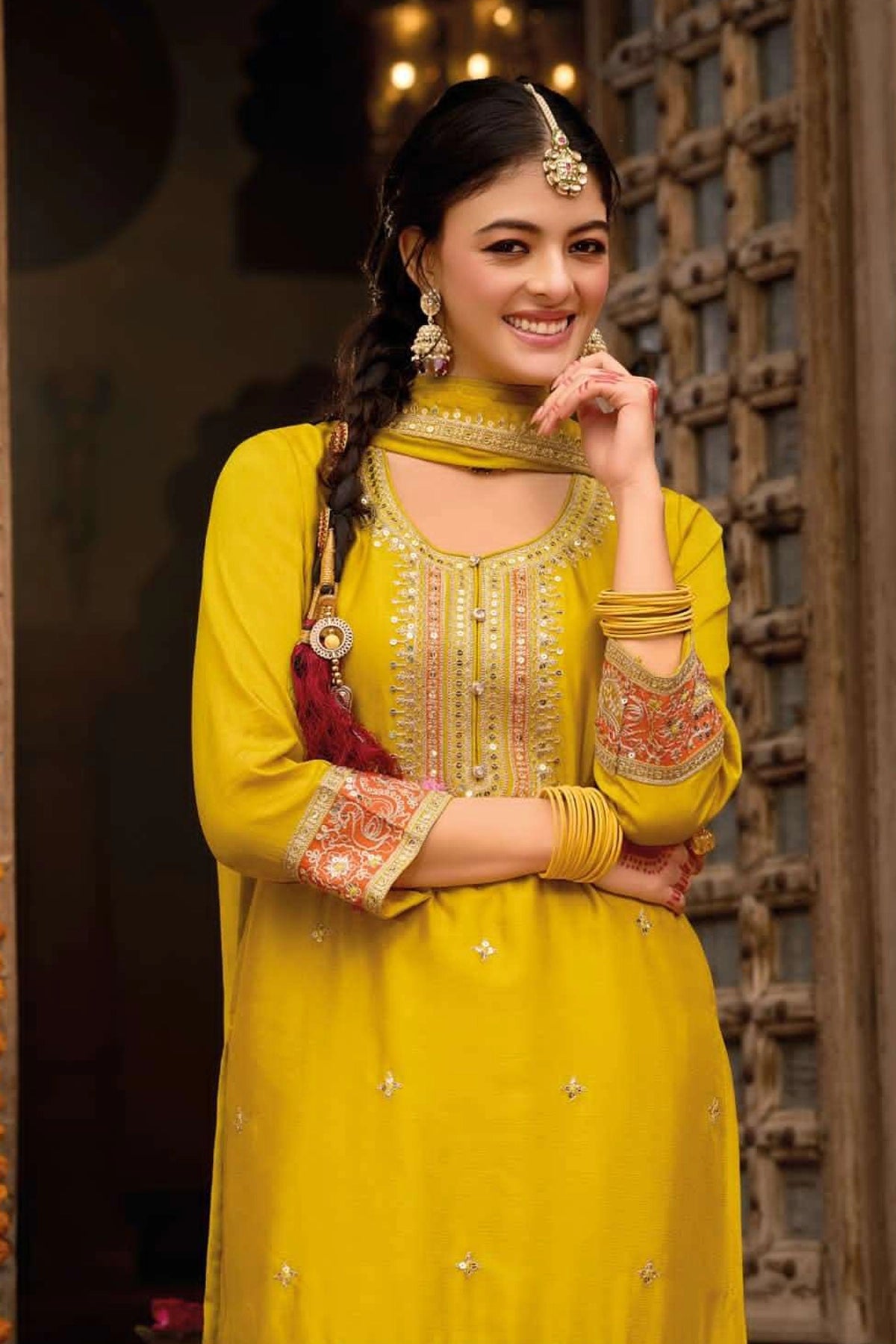 Woman in a yellow traditional punjabi outfit with intricate designs, standing against a decorative background.