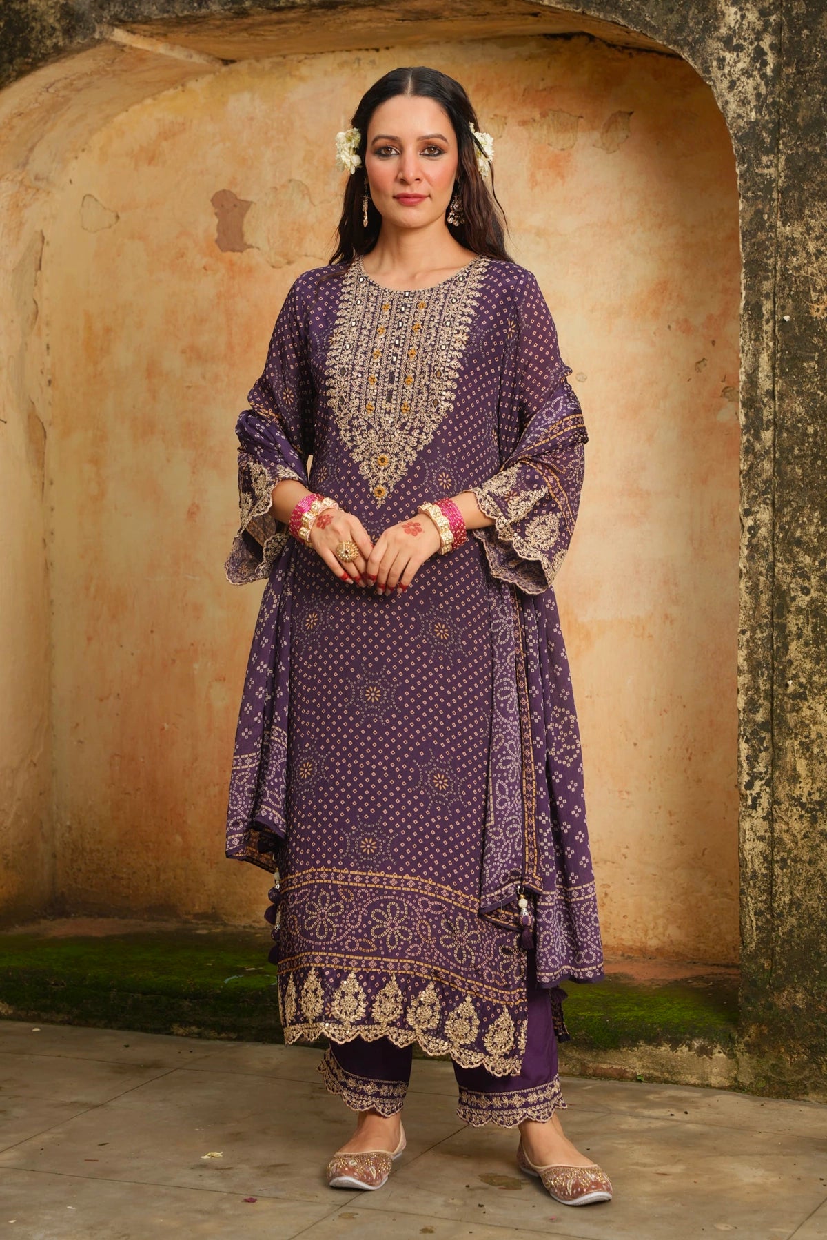 Woman in a traditional purple embroidered outfit standing against a textured wall.