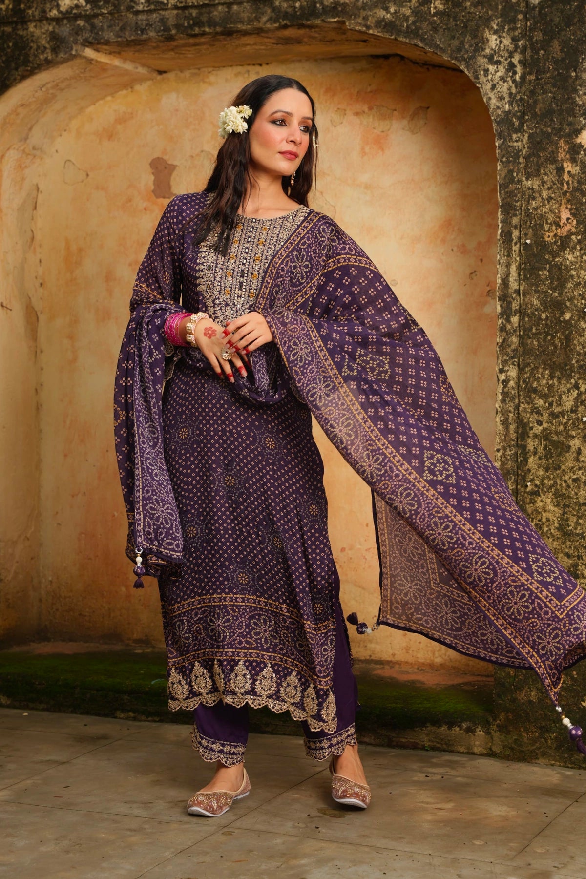 Woman in traditional purple bandhani kurta set with floral headpiece against a textured wall.