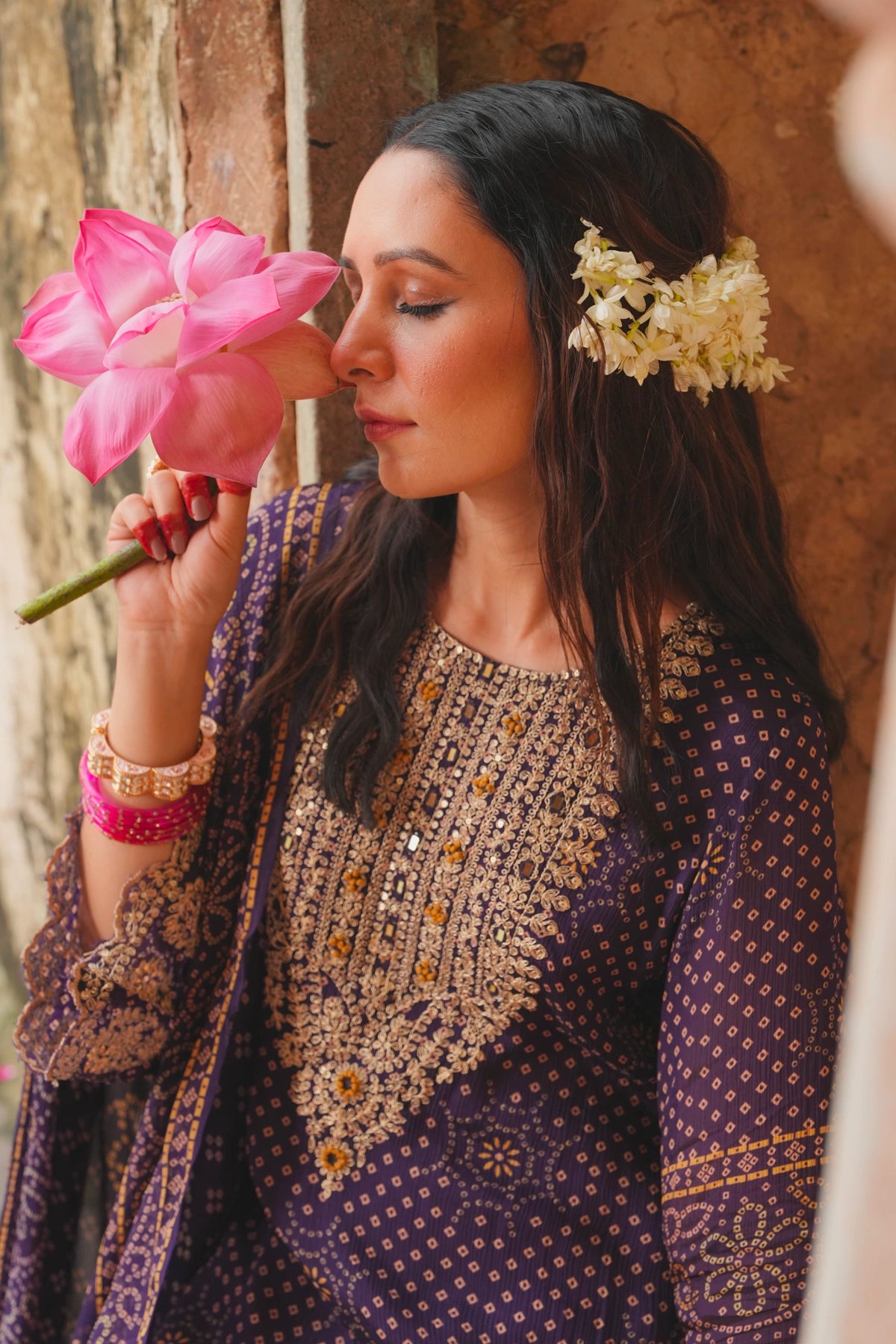 Woman in traditional kurta set attire holding a pink flower against a textured wall.