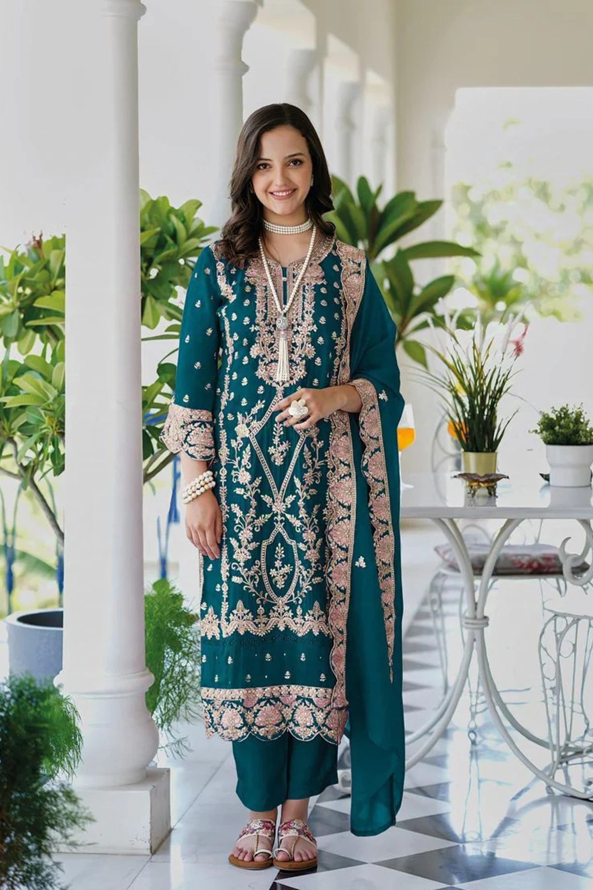 Woman in a teal and pink traditional outfit standing in an elegant indoor setting with plants.