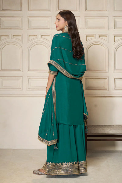 Woman in a teal sharara suit  traditional outfit with gold embroidery against a beige wall.Woman in a teal sharara suit  traditional outfit with gold embroidery against a beige wall.