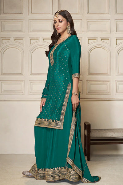 Woman wearing a teal sharara suit traditional outfit with gold embroidery against a beige wall.Woman wearing a teal sharara suit traditional outfit with gold embroidery against a beige wall.