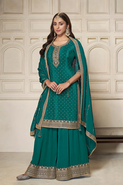 Woman wearing a teal sharara suit traditional outfit with gold embroidery against a beige wall.