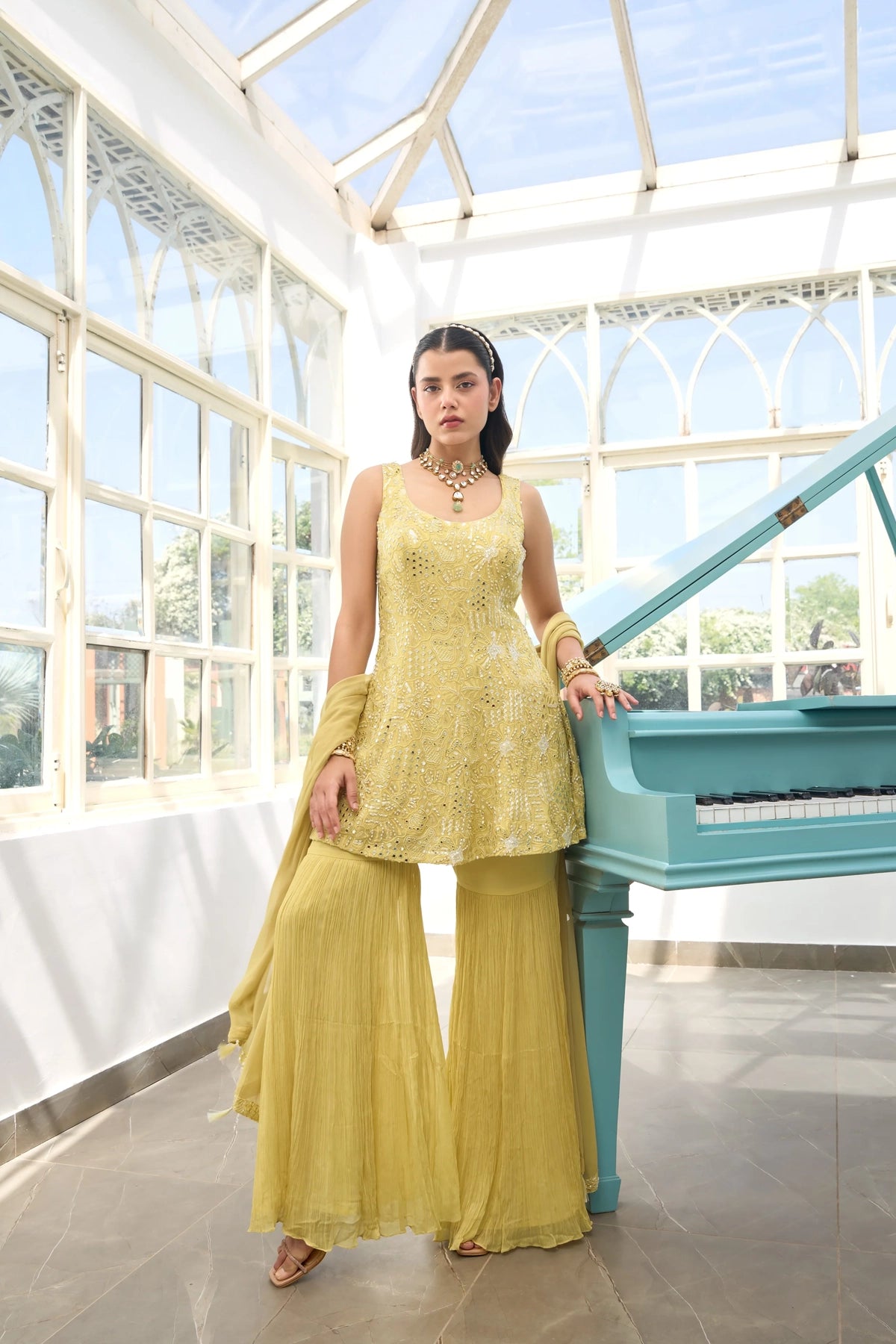 Woman in a Light Yellow Georgette Sharara Suit outfit standing next to a blue piano in a sunlit room with large windows.