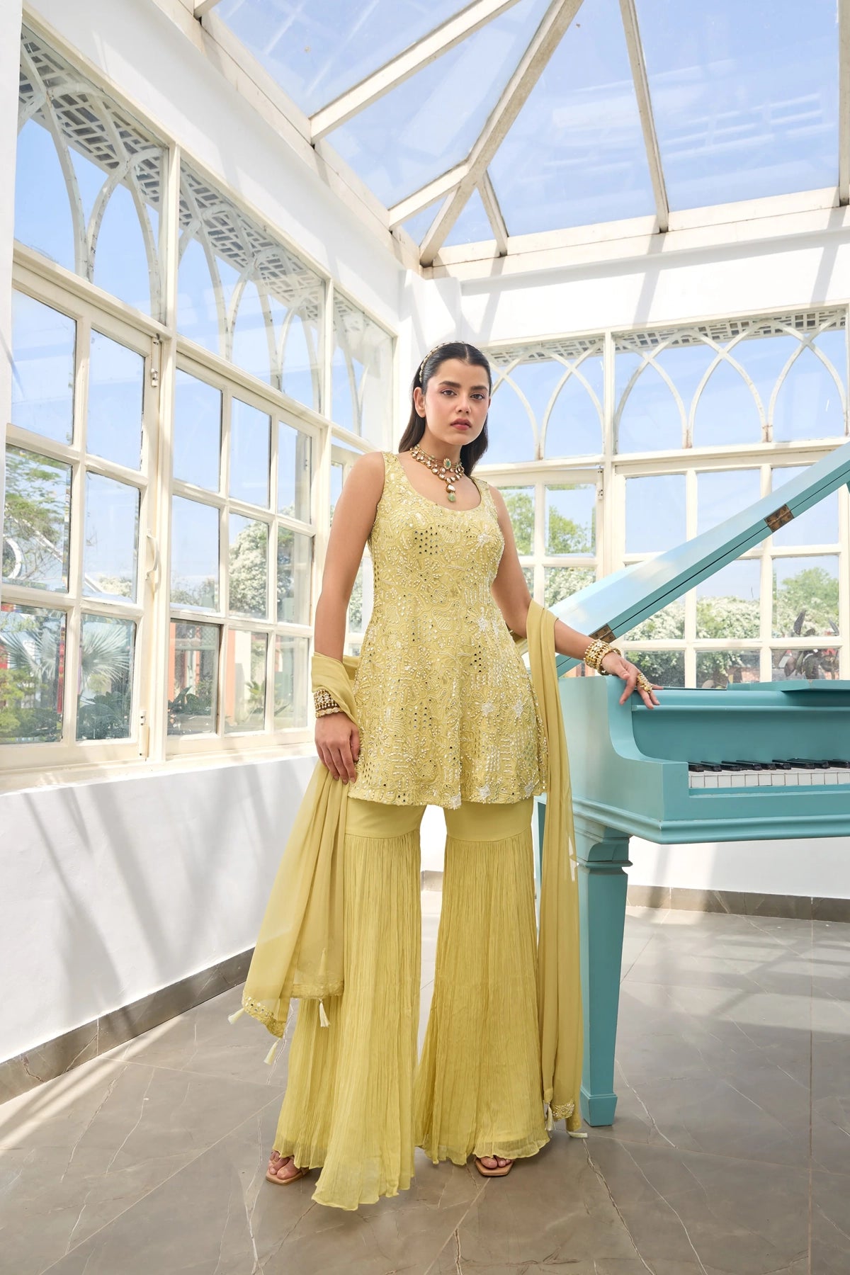 Woman in a Light Yellow Georgette Sharara Suit traditional outfit standing next to a blue piano in a conservatory.